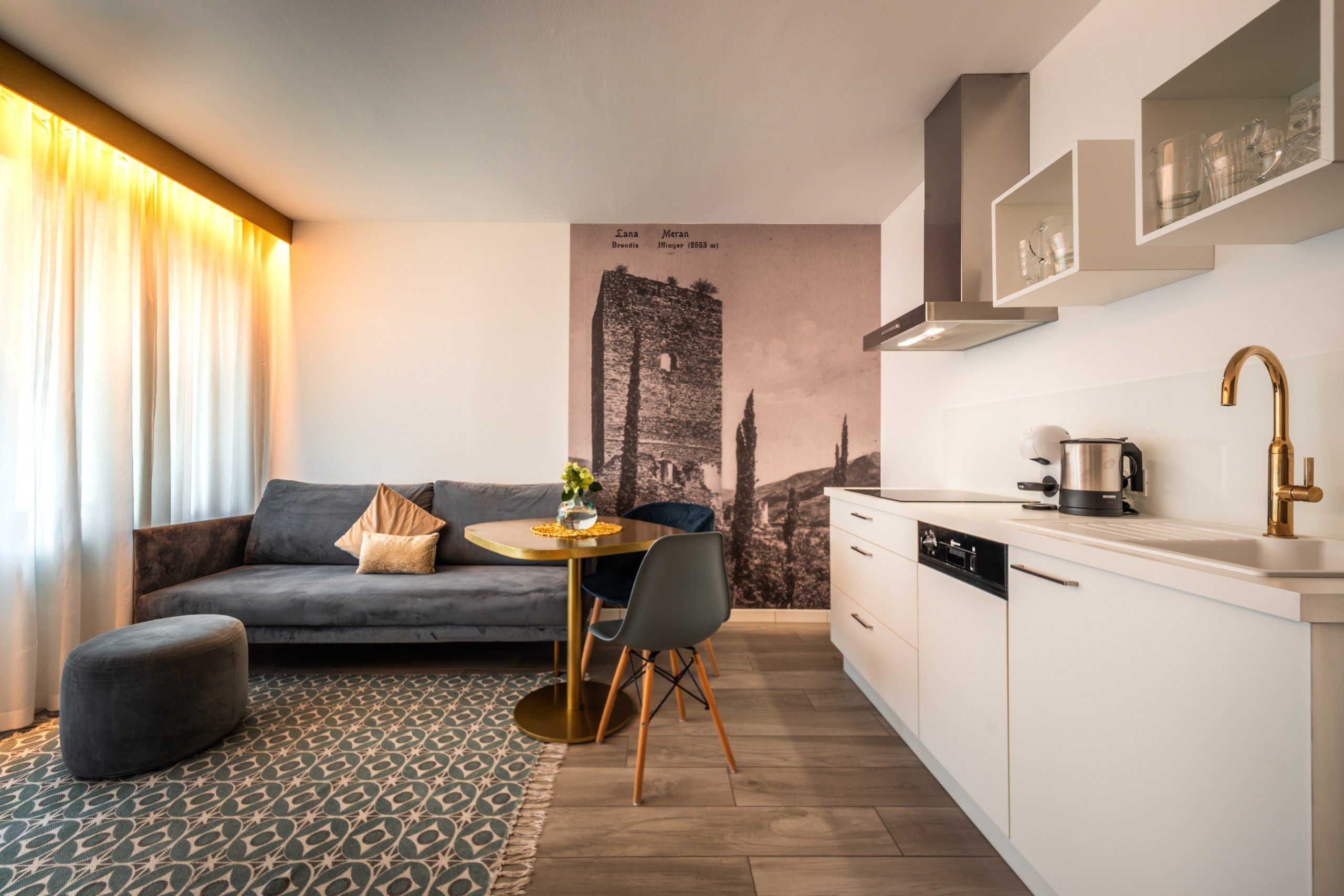 Una cucina moderna con mobili bianchi, un rubinetto dorato e un'area pranzo con un divano grigio e un tavolino rotondo.
