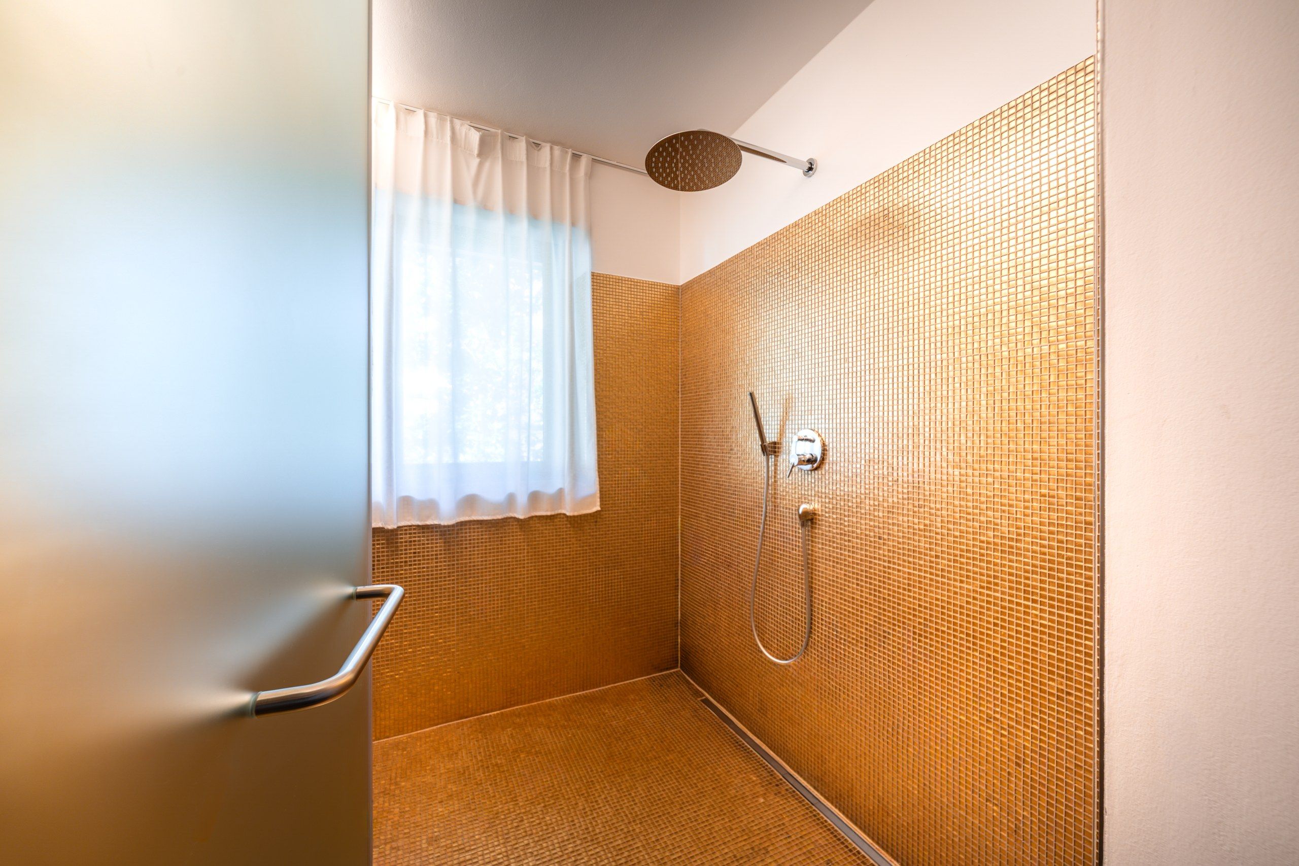 Een douche met een structuurwand, een raam met een wit gordijn en een douchekop.