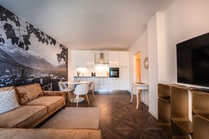 A modern living room with a sofa, a dining area with chairs, a kitchen with cabinets and appliances, and a television.