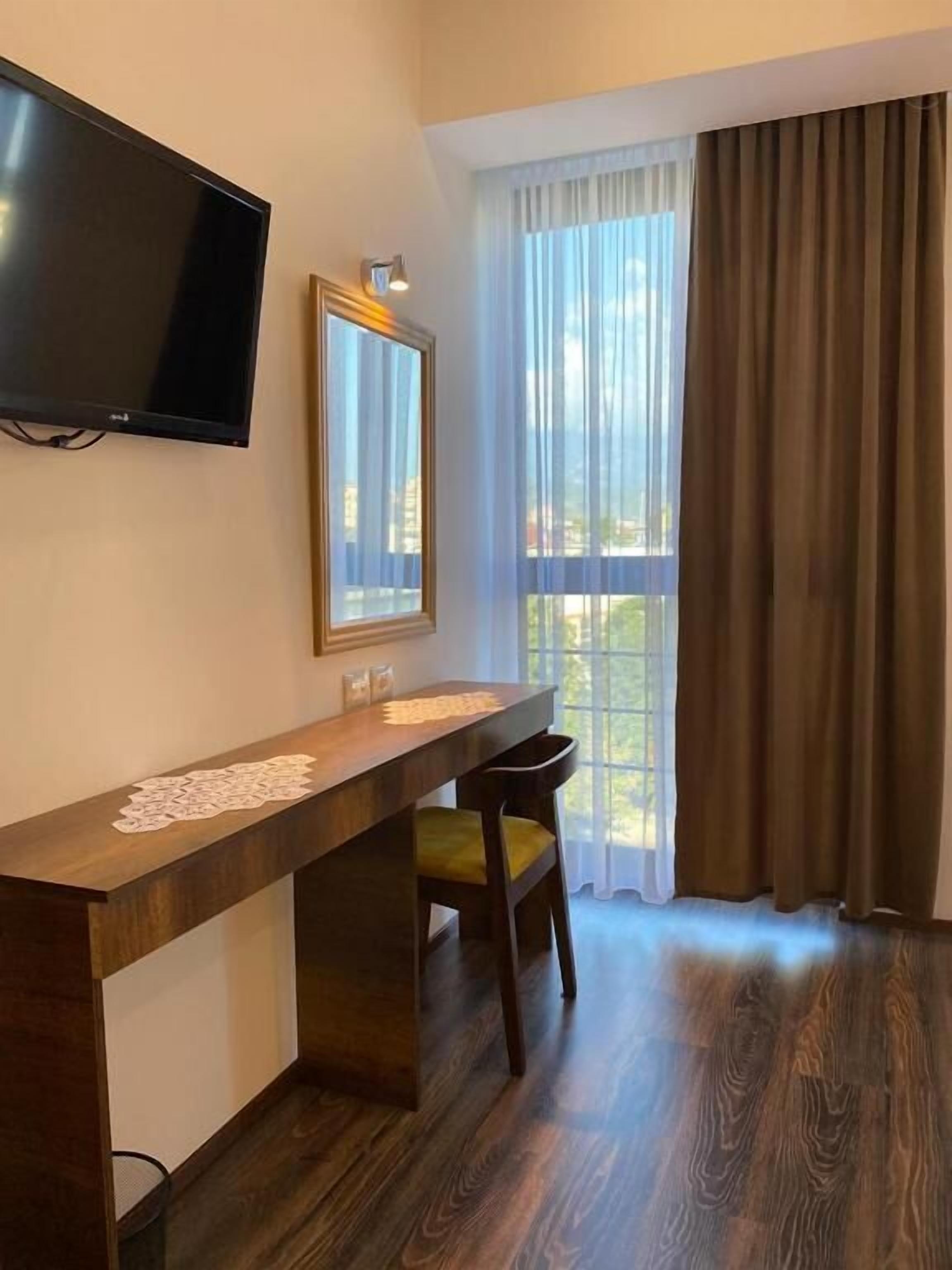 A hotel room with a wooden desk, a flat-screen TV, a large window with curtains, and a view of the outdoors.