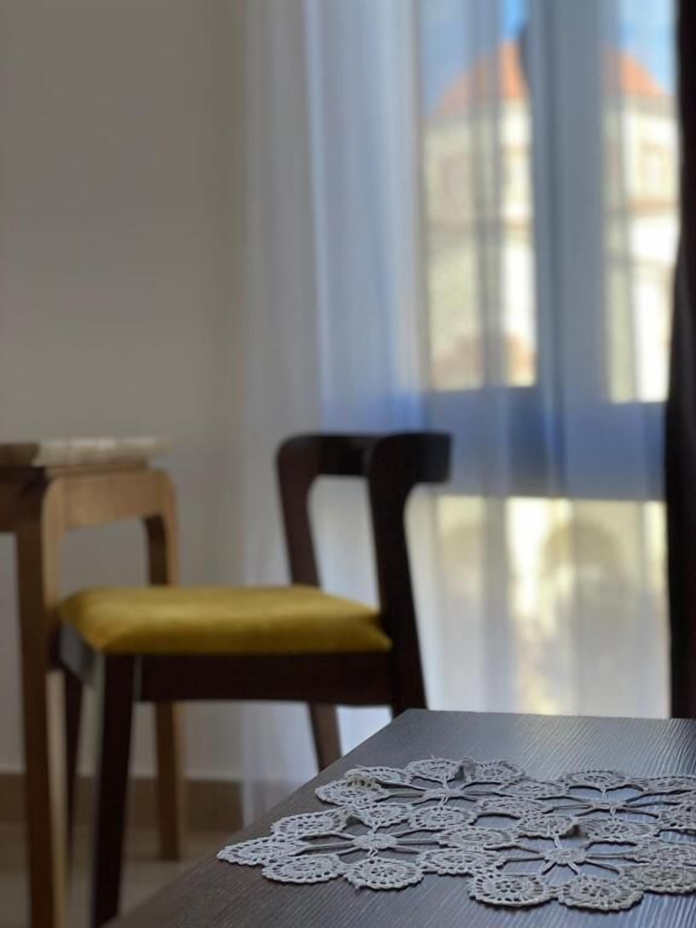 A dining table with a lace table runner, a wooden chair with a yellow cushion, and a window with sheer curtains.