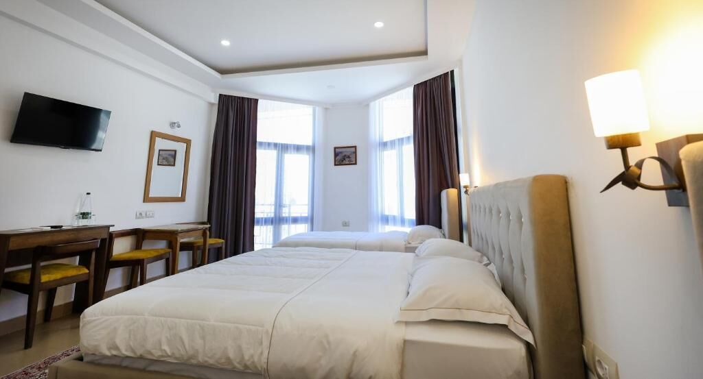 Habitación de hotel con dos camas, un escritorio, sillas, un televisor y una ventana con cortinas.