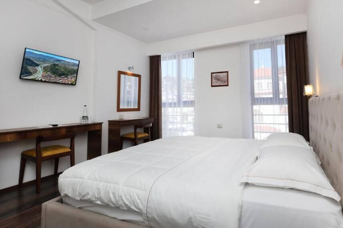 Habitación de hotel con una cama grande, un escritorio con silla, un televisor montado en la pared y una ventana con cortinas.