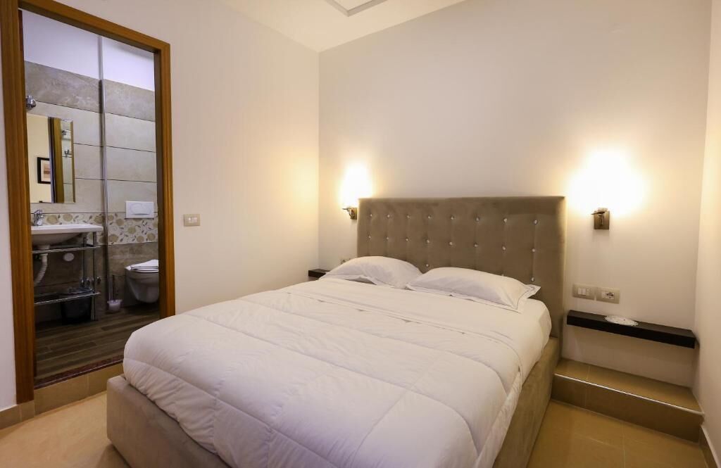 Ein Schlafzimmer mit einem großen Bett, zwei Nachttischlampen, einem hölzernen Kopfteil und einem durch eine Schiebetür einsehbaren Badezimmer.