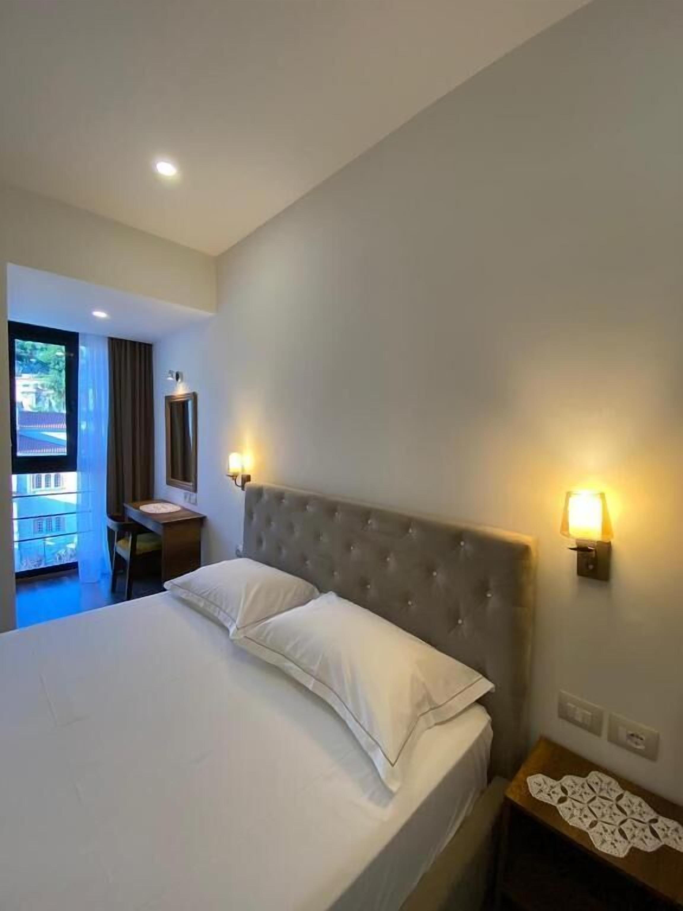 A hotel room with a large bed, a desk, and a chair. There is a window with curtains, a wall-mounted lamp, and a small table with a decorative item.
