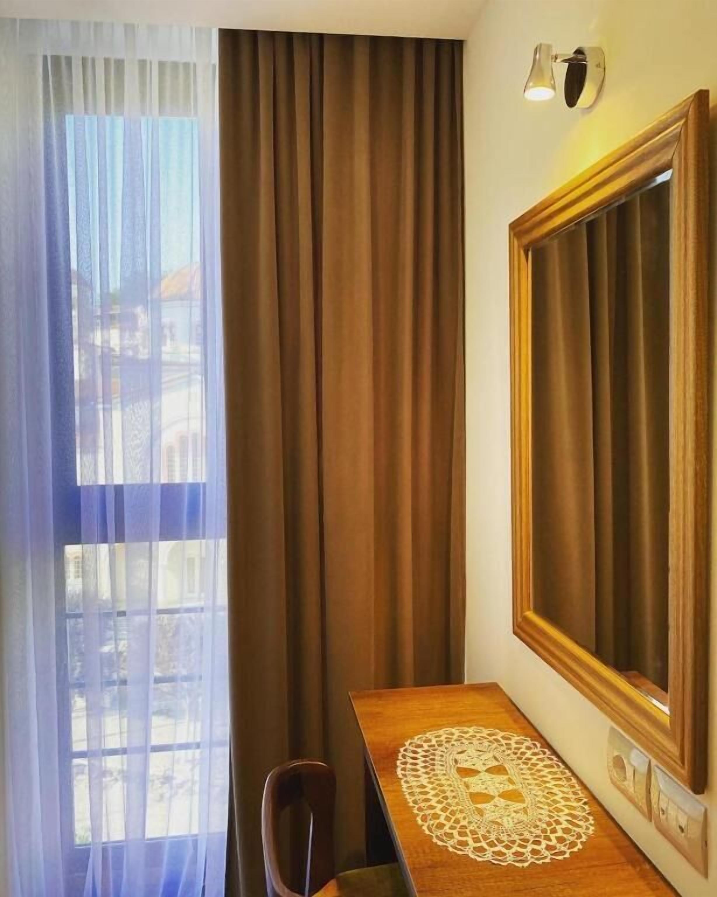 A hotel room with a wooden desk, a chair, a large mirror, and a window with sheer curtains.
