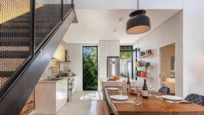 A modern kitchen with a central island, a dining area with a wooden table, and a staircase with a metal railing. - Views, Fireplace, Sea Point - Mnt Nelson Townhouse (Cape Town)