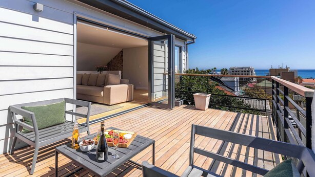 A balcony with a wooden deck, a sofa, and a table with a bottle and glasses. - Views, Fireplace, Sea Point - Mnt Nelson Townhouse (Cape Town)
