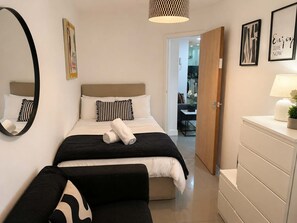 A hotel room with a bed, a bedside table with a lamp, a mirror, and a door leading to a bathroom. - 2 BR - Fast Wifi - Near the City Centre (Cardiff)