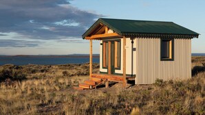 Una cabaña de madera pequeña con techo verde, ubicada en una colina cubierta de hierba con vista a un cuerpo de agua.