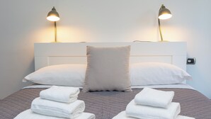 Een net opgemaakt bed met witte kussens en opgevouwen handdoeken, twee bedlampen en een modern hoofdeinde.