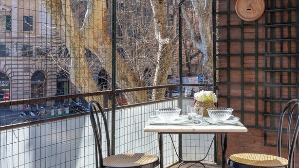 Een balkon met een tafel voor twee, stoelen en een uitzicht op een stadslandschap met gebouwen en een boom.