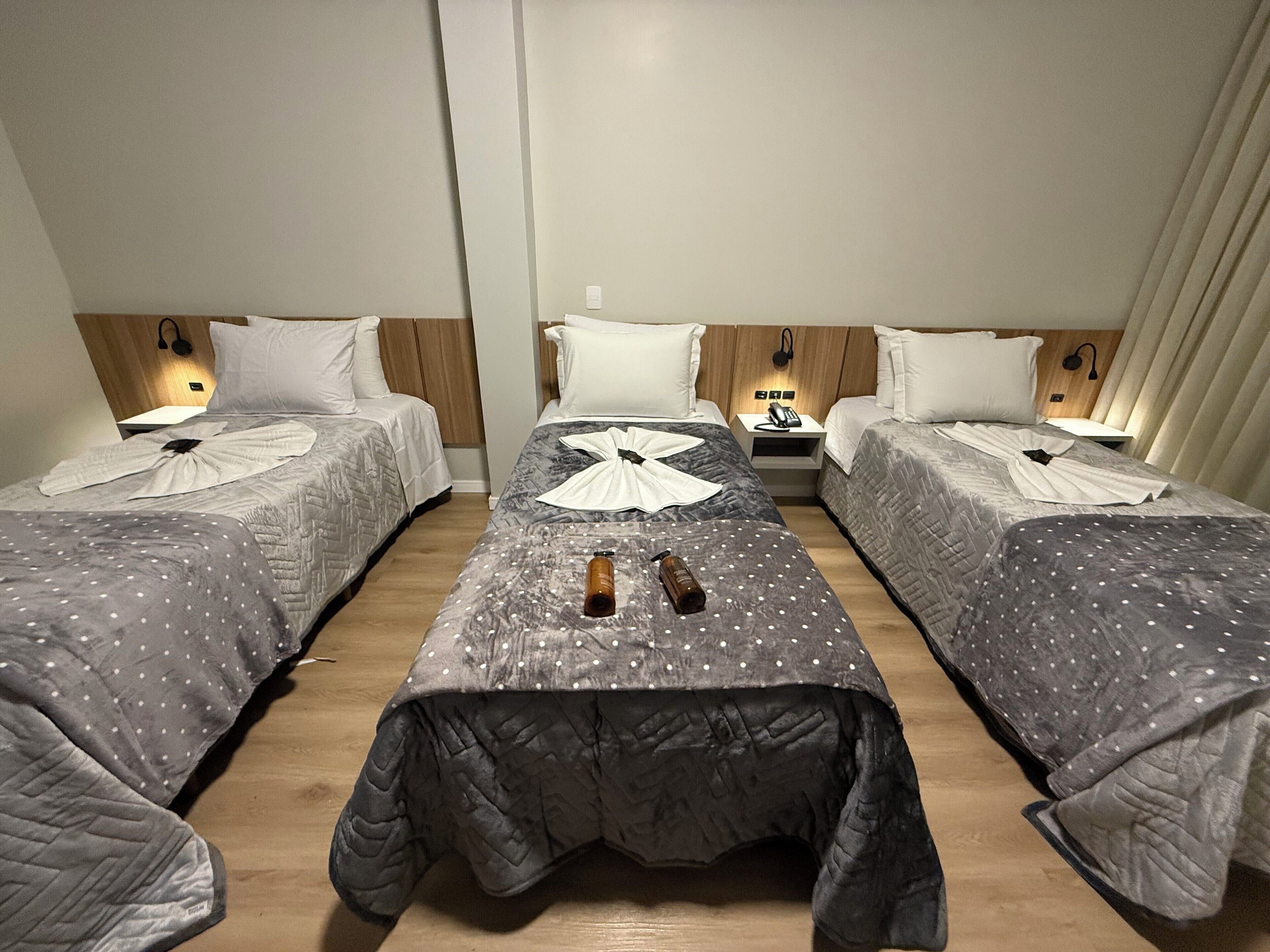 Habitación de hotel con tres camas individuales, cada una con cabecera de madera, almohadas blancas y una mesita de noche con lámpara.