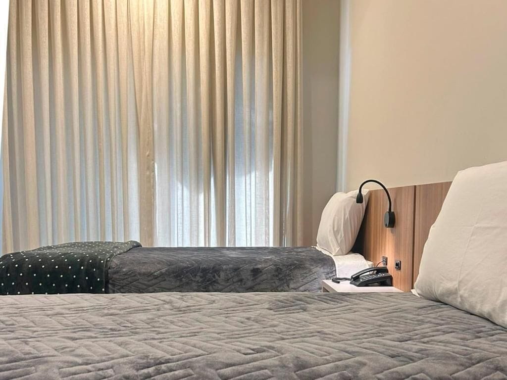 A hotel room with a bed, a bedside table with a lamp, and a window with curtains.