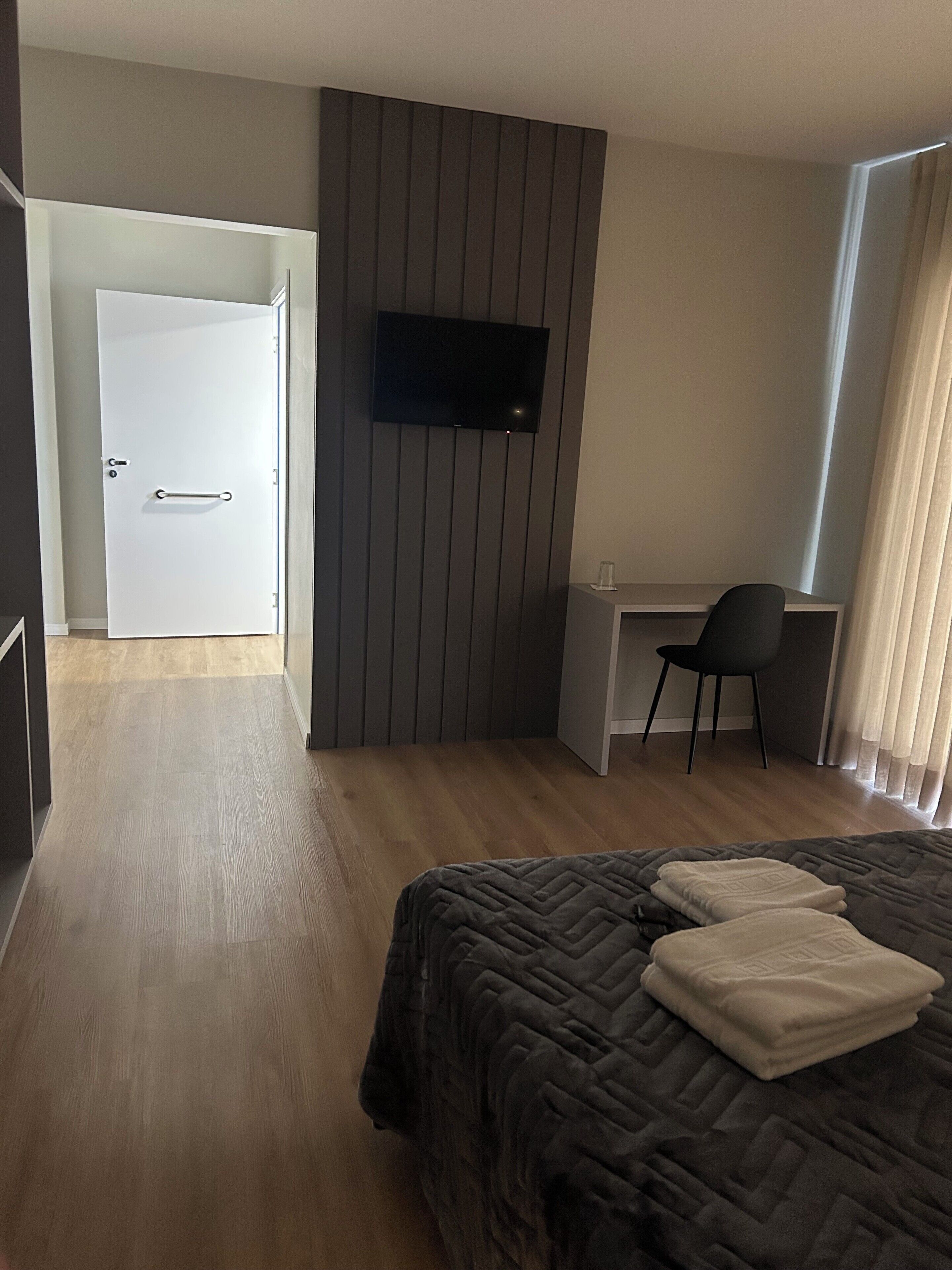 A hotel room with a bed, a desk, a chair, a TV mounted on the wall, and a door leading to another room.