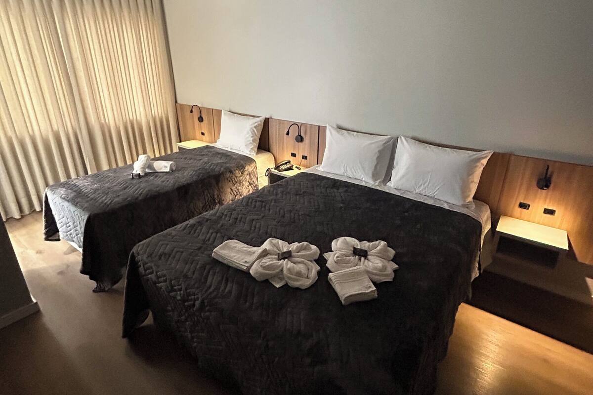 A hotel room with two beds, a wooden headboard, bedside lamps, and a large window with curtains.