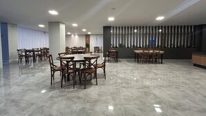 A spacious conference room with multiple tables and chairs, a modern ceiling with recessed lighting, and a large window with vertical blinds. - Bon Vivant Hotel (Fraiburgo)