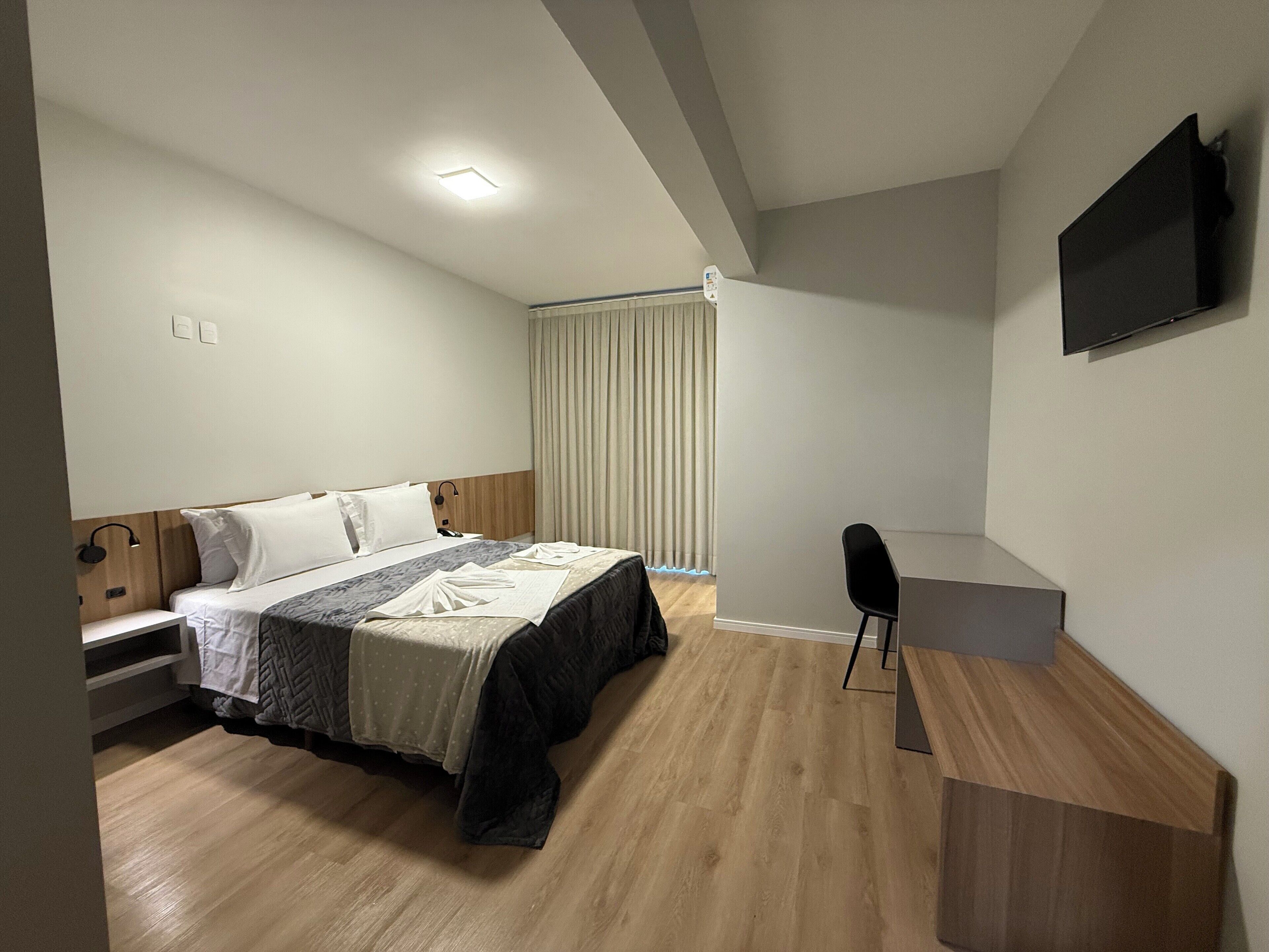 A modern hotel room with a bed, bedside table, desk, and a flat-screen TV mounted on the wall.