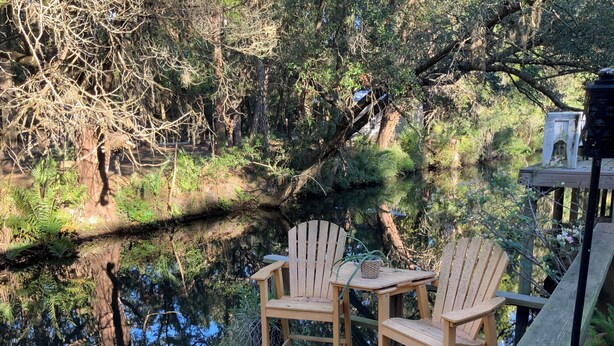 Una terraza de madera con dos sillas Adirondack que dan a una masa de agua tranquila, rodeada de abundante follaje.