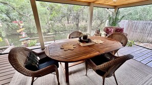 An outdoor dining area with a wooden table and wicker chairs, surrounded by a wooden deck and a view of a pond.