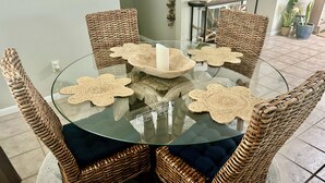 Una mesa redonda de vidrio con un cuenco en el centro, rodeada por cuatro sillas de mimbre con cojines oscuros.
