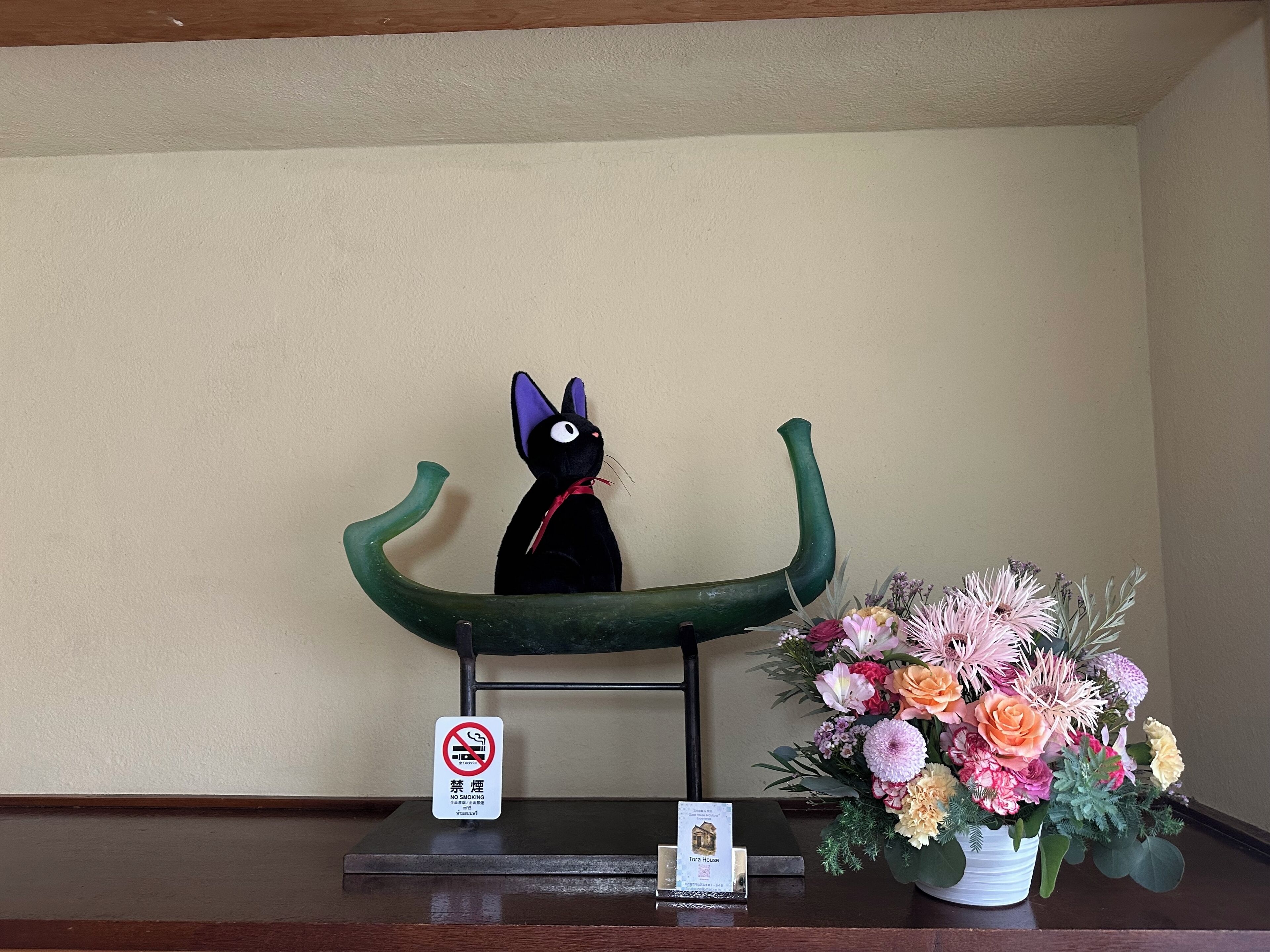 Una statuina di un gatto nero su una poltrona verde, un cartello di divieto di fumare e un bouquet di fiori su un tavolo.