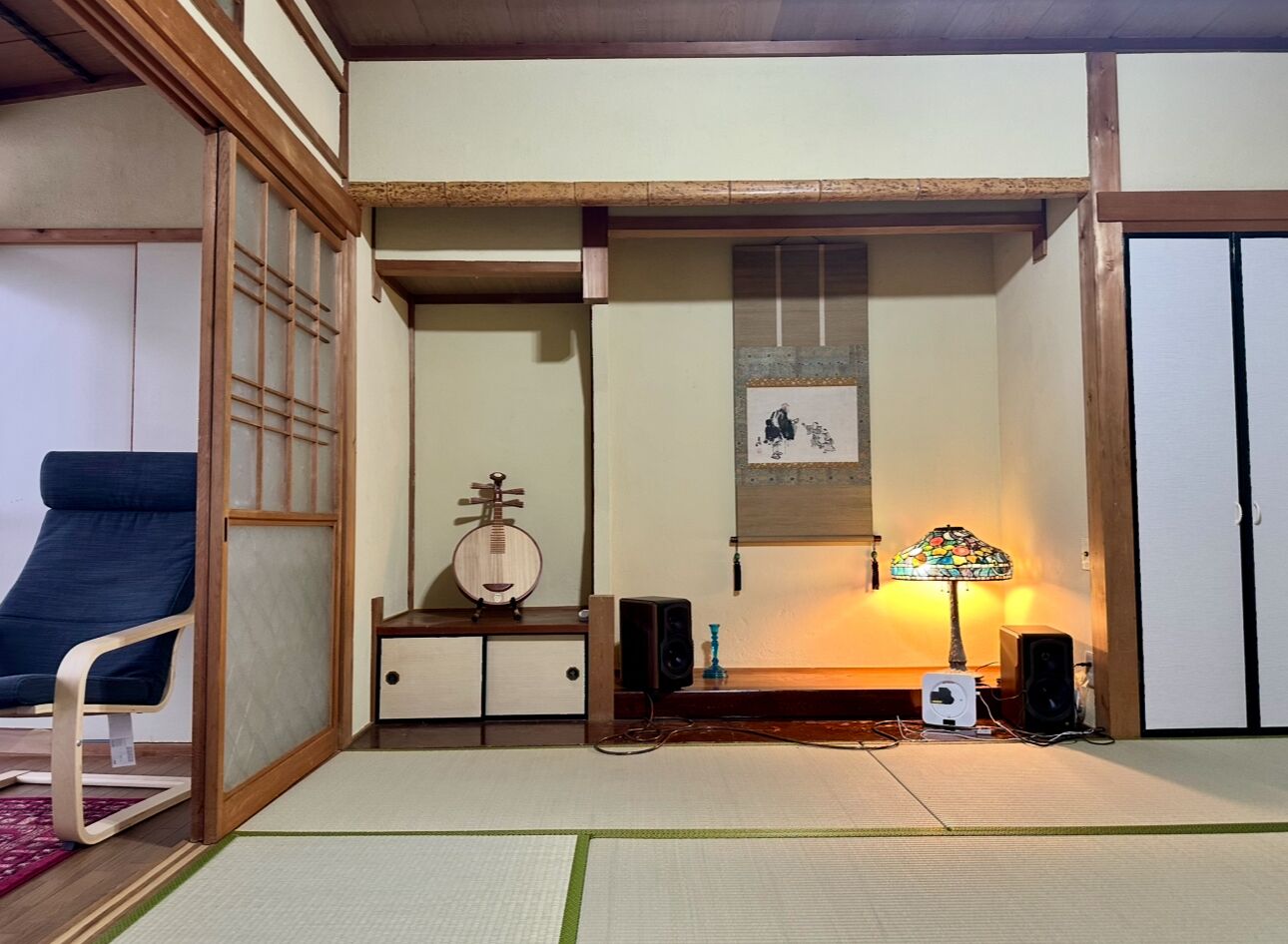 Una tradizionale stanza giapponese con pavimenti in tatami, un tavolino con un oggetto decorativo, una sedia, un altoparlante a parete e una lampada.