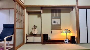 Una tradizionale stanza giapponese con pavimenti in tatami, un tavolino con un oggetto decorativo, una sedia, un altoparlante a parete e una lampada.