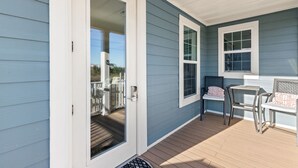 A covered porch with a sliding glass door, a small table, two chairs, and a window with a view of the deck. - Beach House - C Shucker by PHG (Mexico Beach)