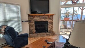 A living room with a stone fireplace, a flat-screen TV mounted above it, a blue armchair, a wooden coffee table, and a lamp. - KLTD1  Klonteska Manor  An unforgettable mountain getaway! (Beech Mountain)