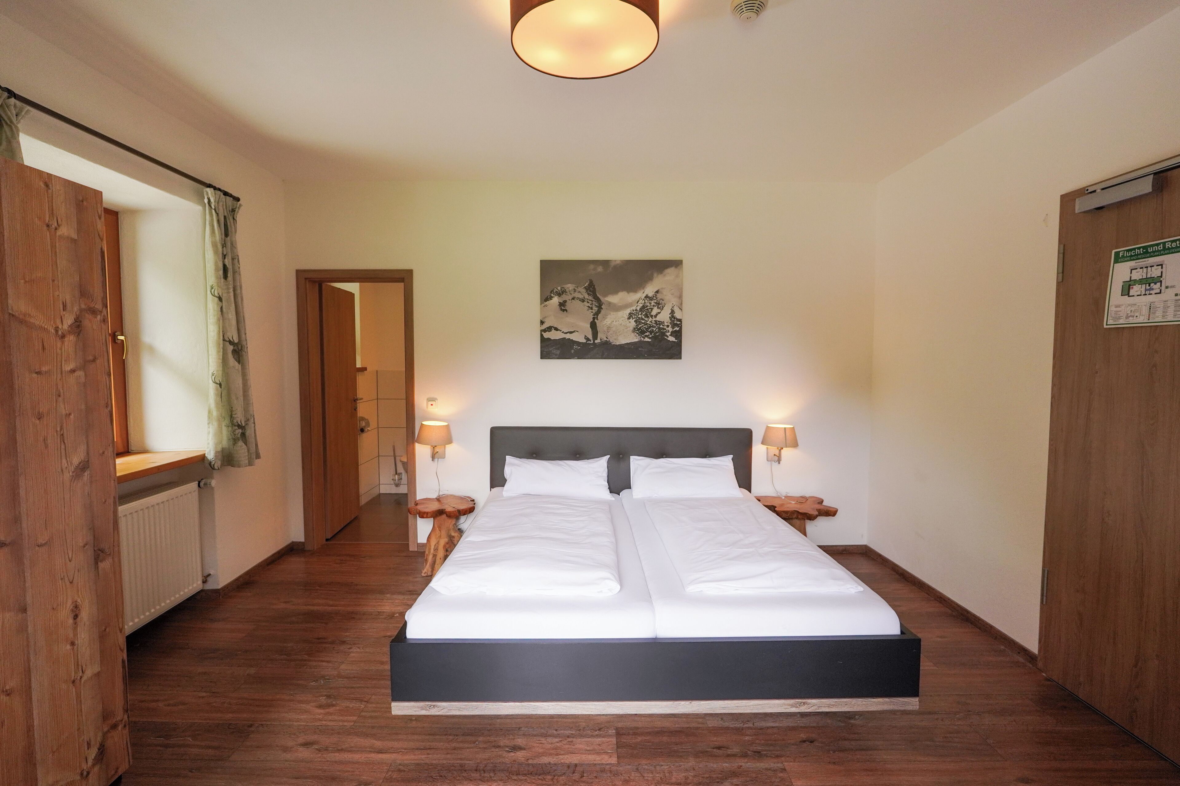 A double bed with white linens, two bedside tables with lamps, a wooden headboard, a wall-mounted painting of a mountain, and a wooden wardrobe.
