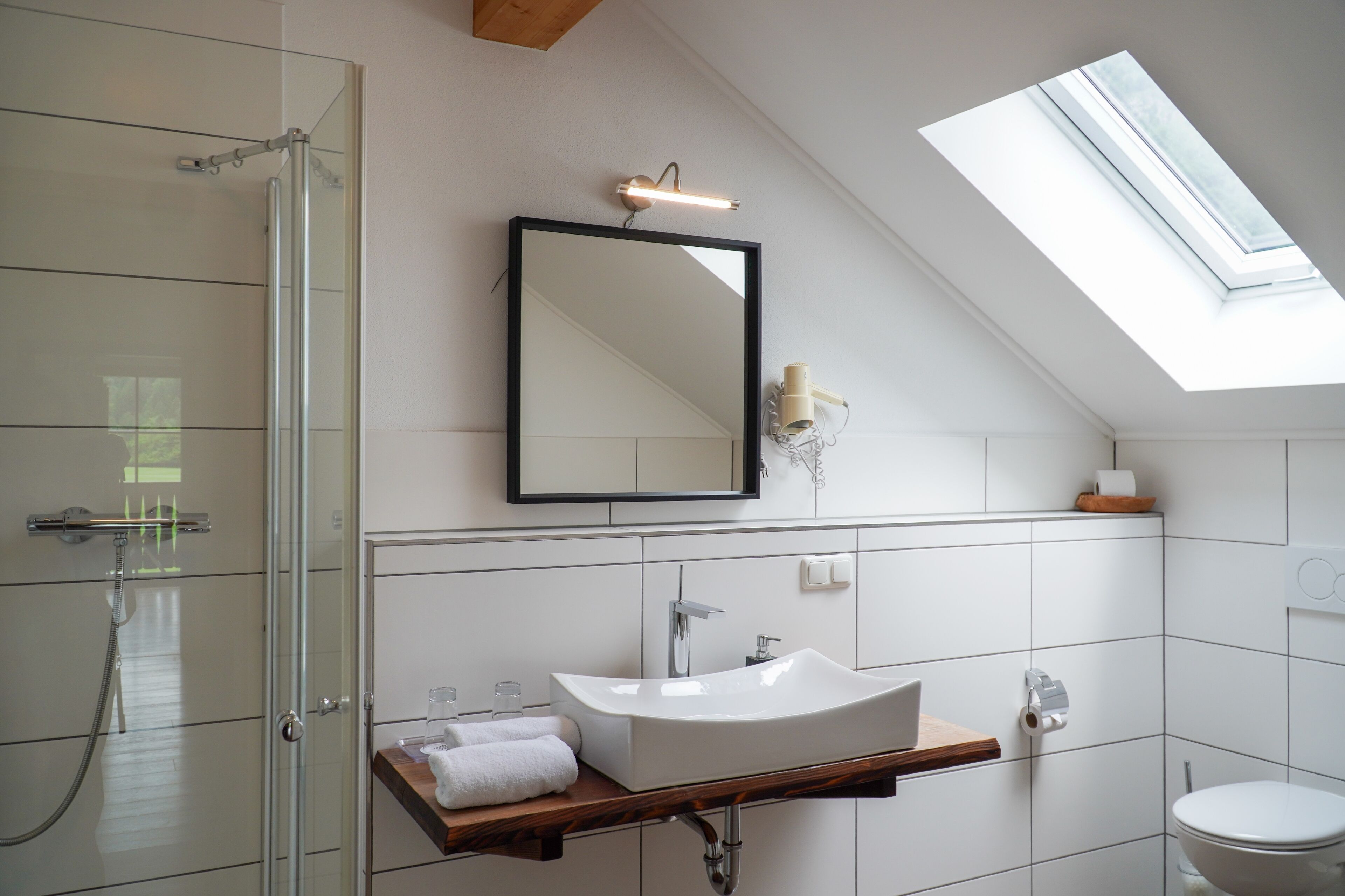 A modern bathroom with a skylight, a glass shower enclosure, a white sink, and a wooden vanity.