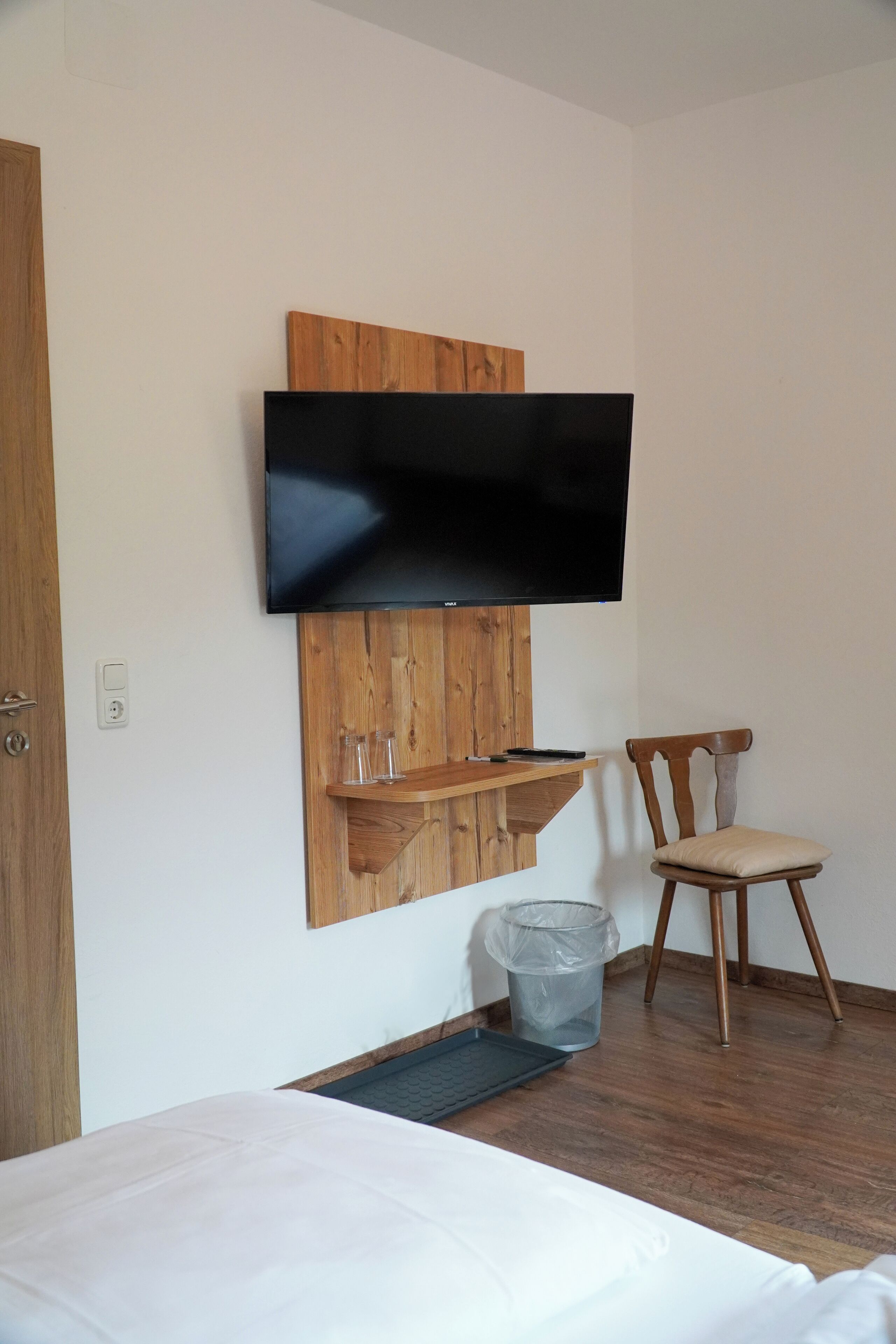 A room with a bed, a wooden shelf with glasses, a chair, and a mounted TV.
