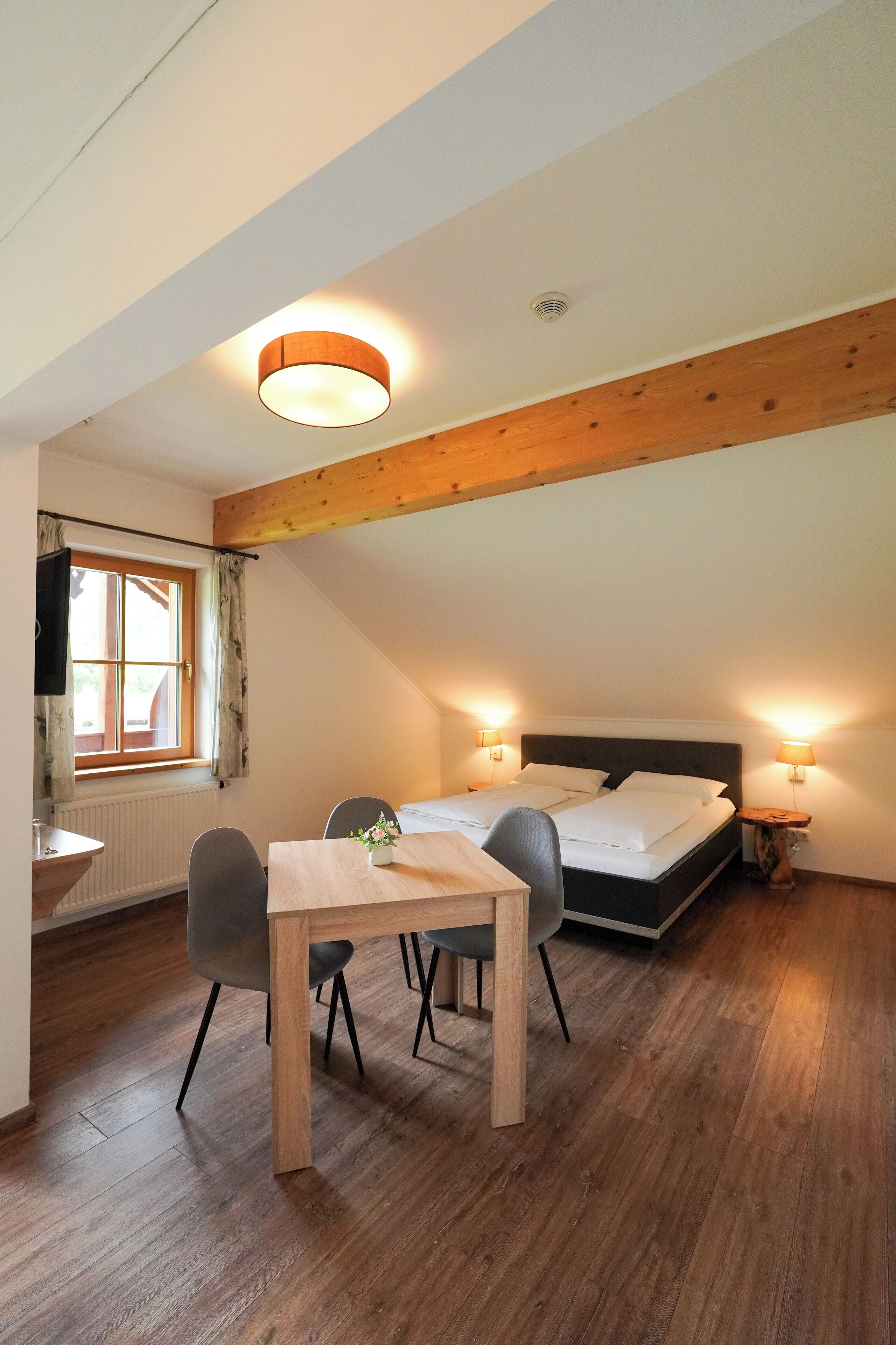 A cozy room with a wooden ceiling, a bed with white bedding, a dining table with chairs, and two bedside lamps.