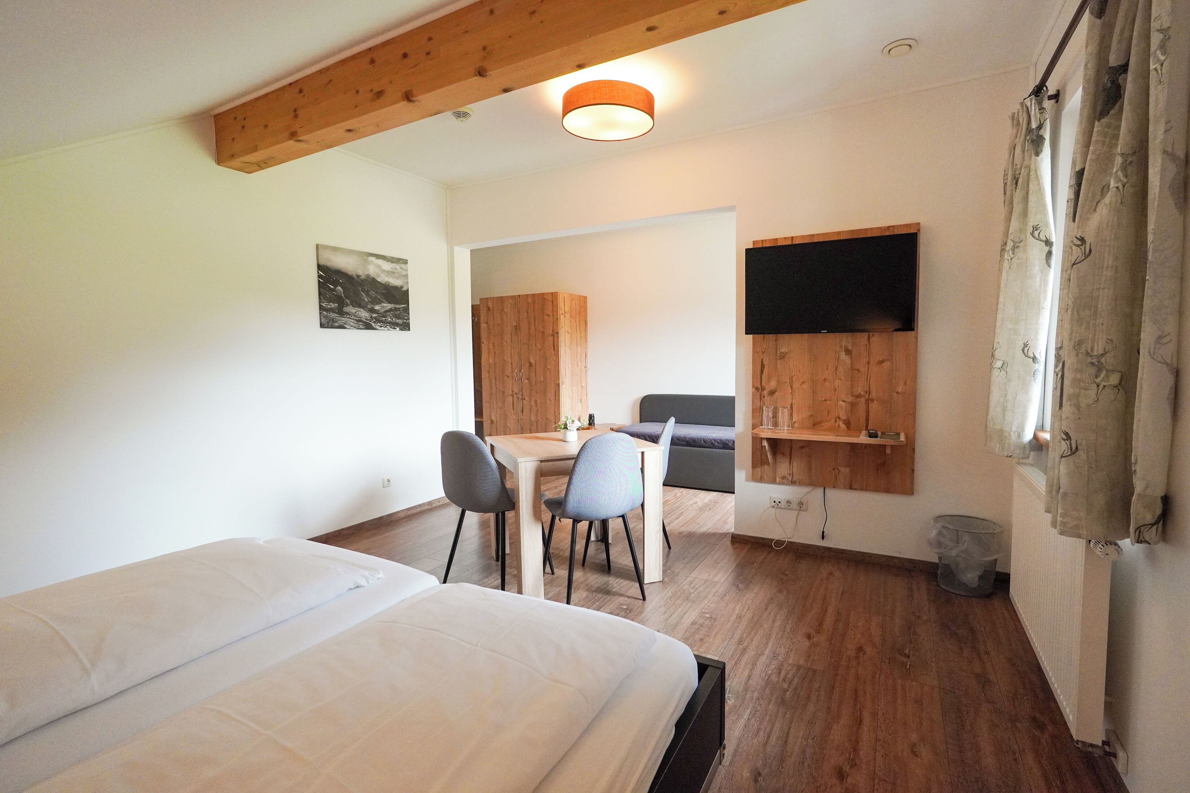 A bedroom with a bed, a desk, and a chair. There is a wooden ceiling beam, a wall-mounted TV, and a window with curtains.