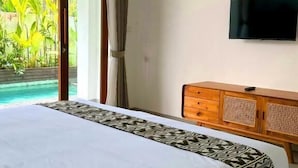 A hotel room with a large bed, wooden headboard, a television mounted on the wall, and a view of a swimming pool and palm trees through the window. - Kemari Ubud (Ubud)
