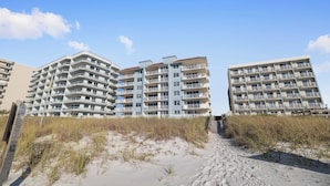 Et hotel ved stranden med flere balkoner, en sandstrand foran og klare himmel.