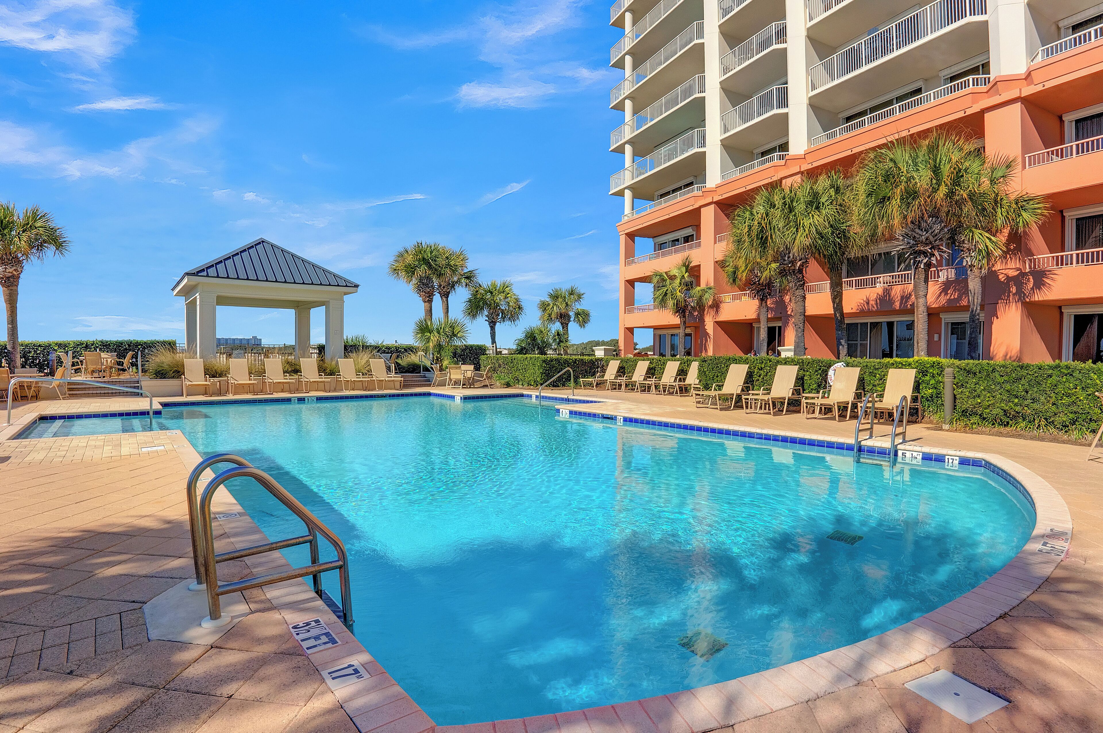 A swimming pool with lounge chairs and a gazebo, surrounded by palm trees and a multi-story building.