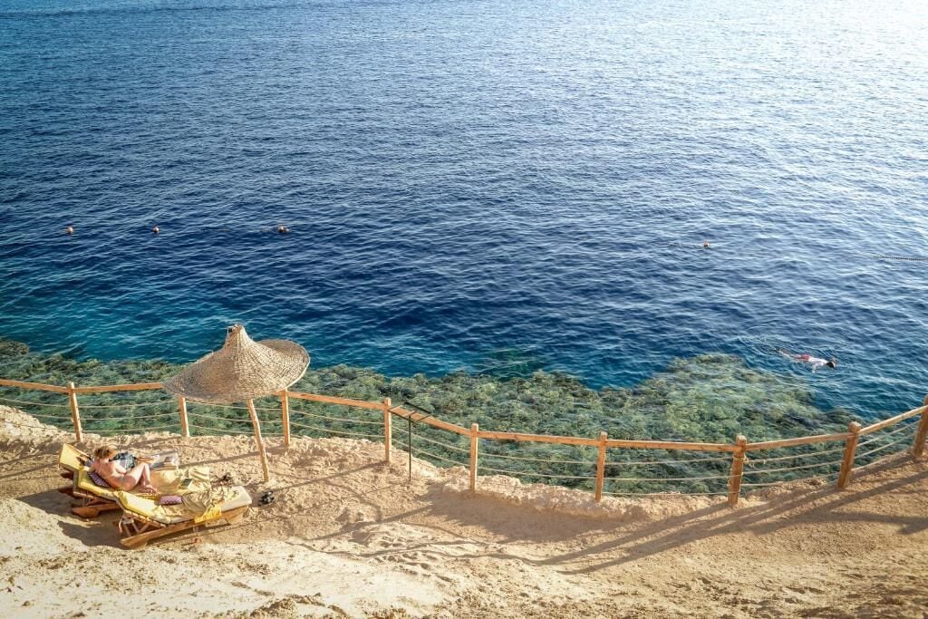 A beach with clear blue water, a wooden fence, and a straw umbrella.