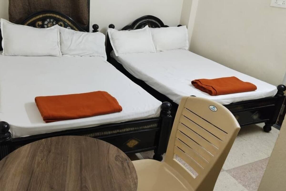 A room with two single beds, each with white linens and an orange blanket. There is a wooden chair and a small round table in the foreground.