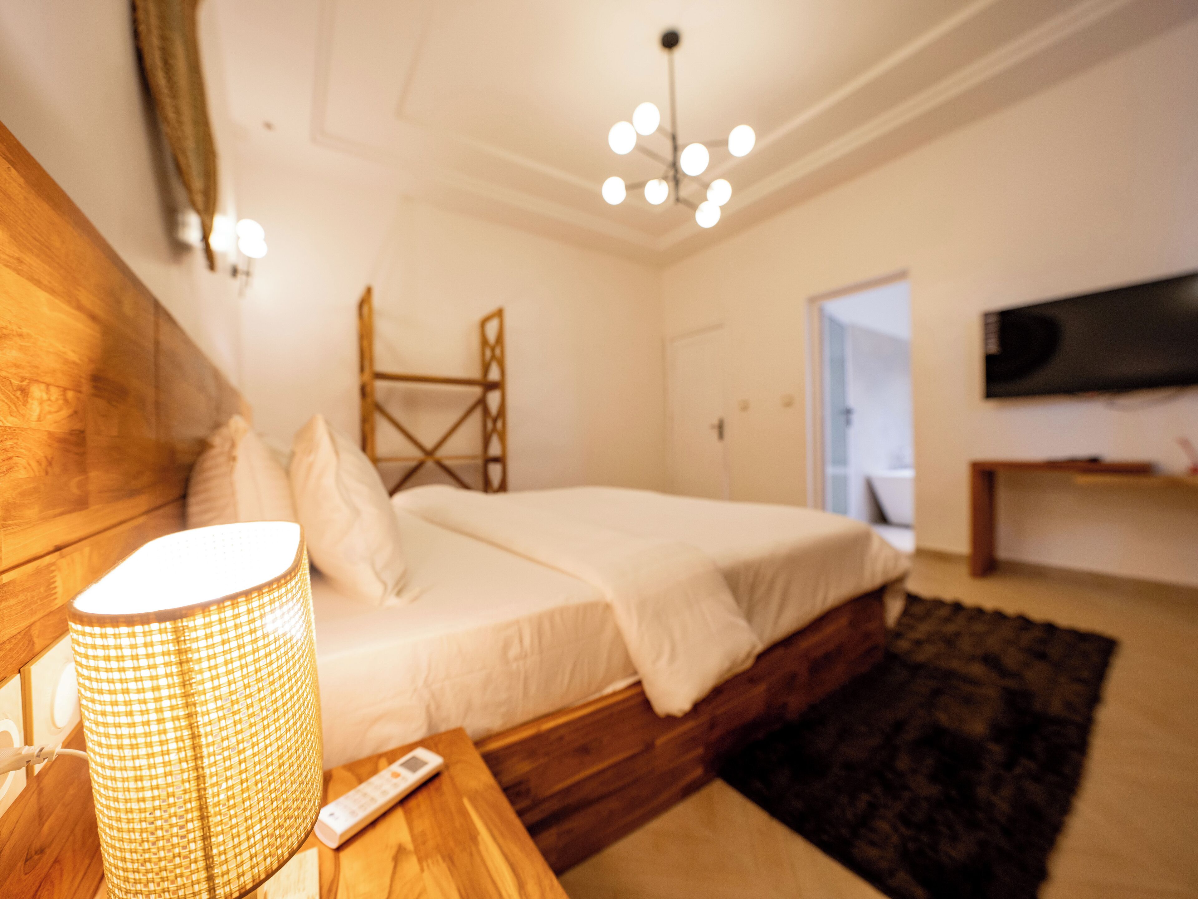 Un dormitorio con una cama, cabecera de madera, mesita de noche con lámpara, control remoto y televisor montado en la pared.