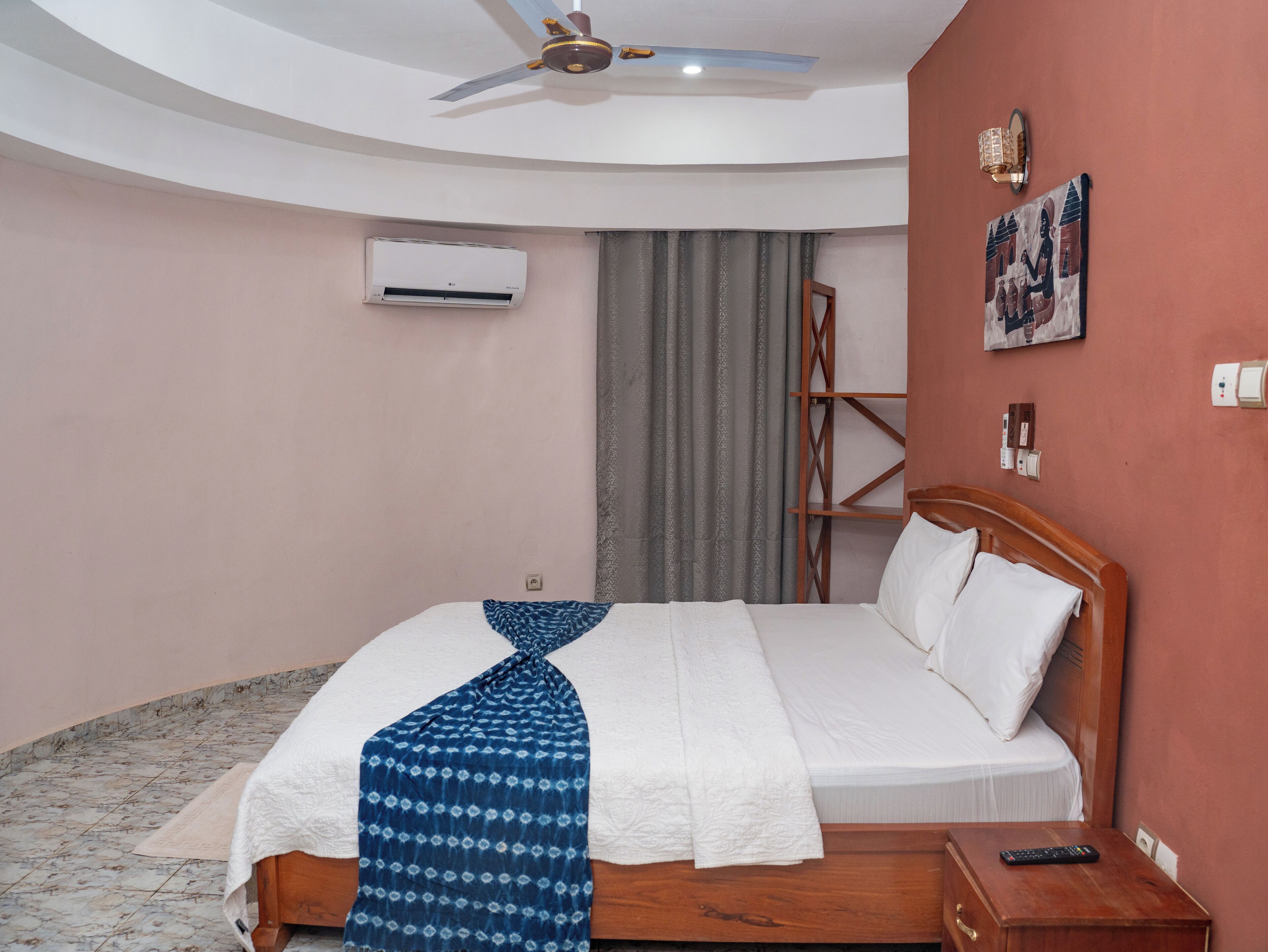 A bedroom with a bed, a ceiling fan, an air conditioning unit, a wooden nightstand, a remote control, and a wall-mounted light fixture.