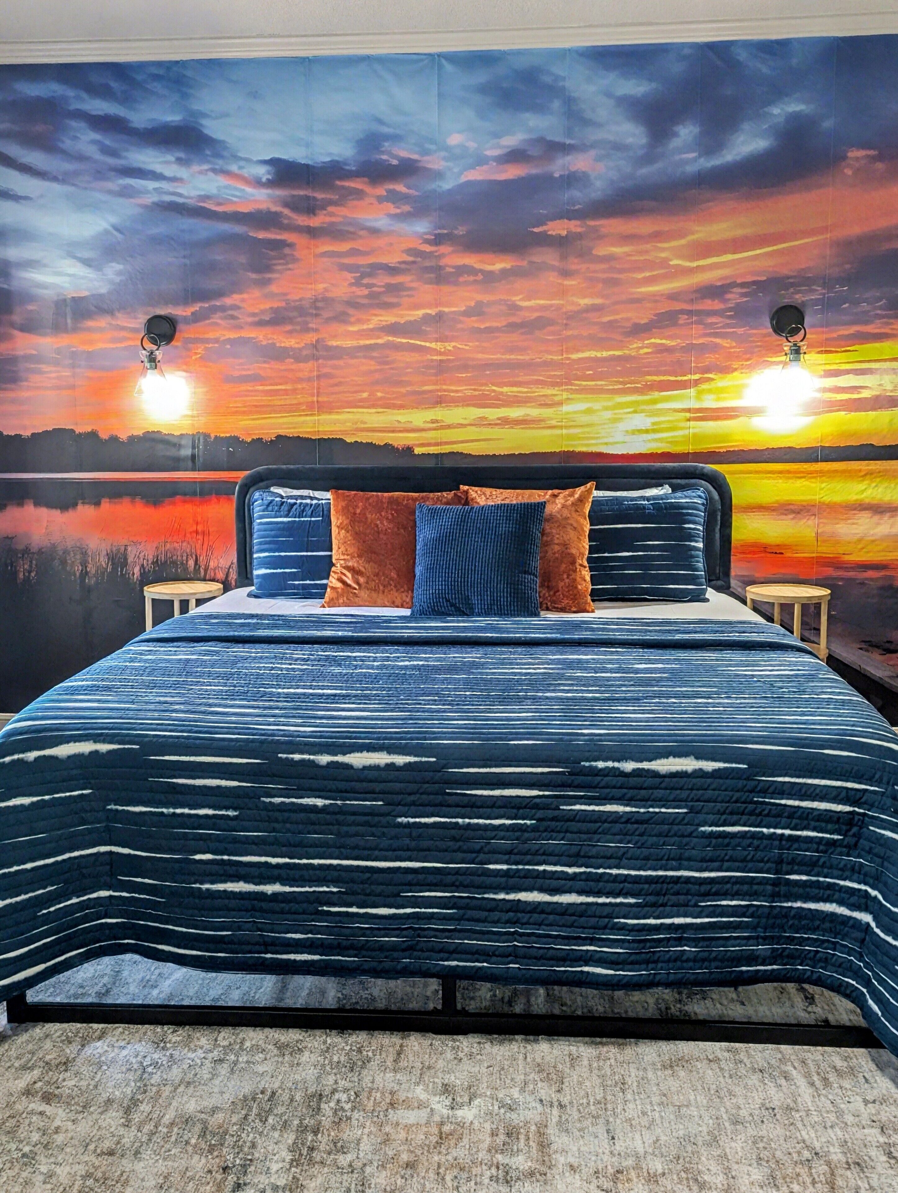 Et soveværelse med en stor seng, udsigt til en solnedgang over vandet, og to lamper på hver side af sengen.