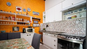 A kitchen with white cabinets, a gas stove, and a microwave. There are shelves with dishes and a dining table with a floral tablecloth. - House w/ Ocean View, Pool and Beach Access Ize0016 (Florianópolis)