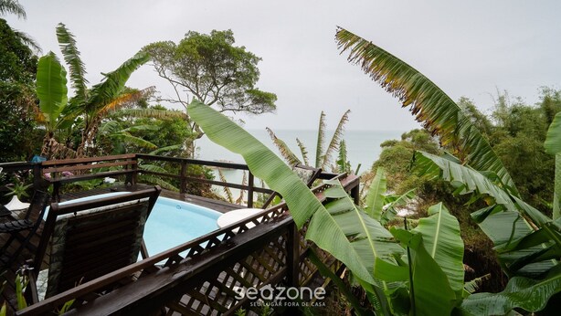 A pool area with wooden deck and lounge chairs, surrounded by tropical foliage and overlooking a body of water. - House w/ Ocean View, Pool and Beach Access Ize0016 (Florianópolis)