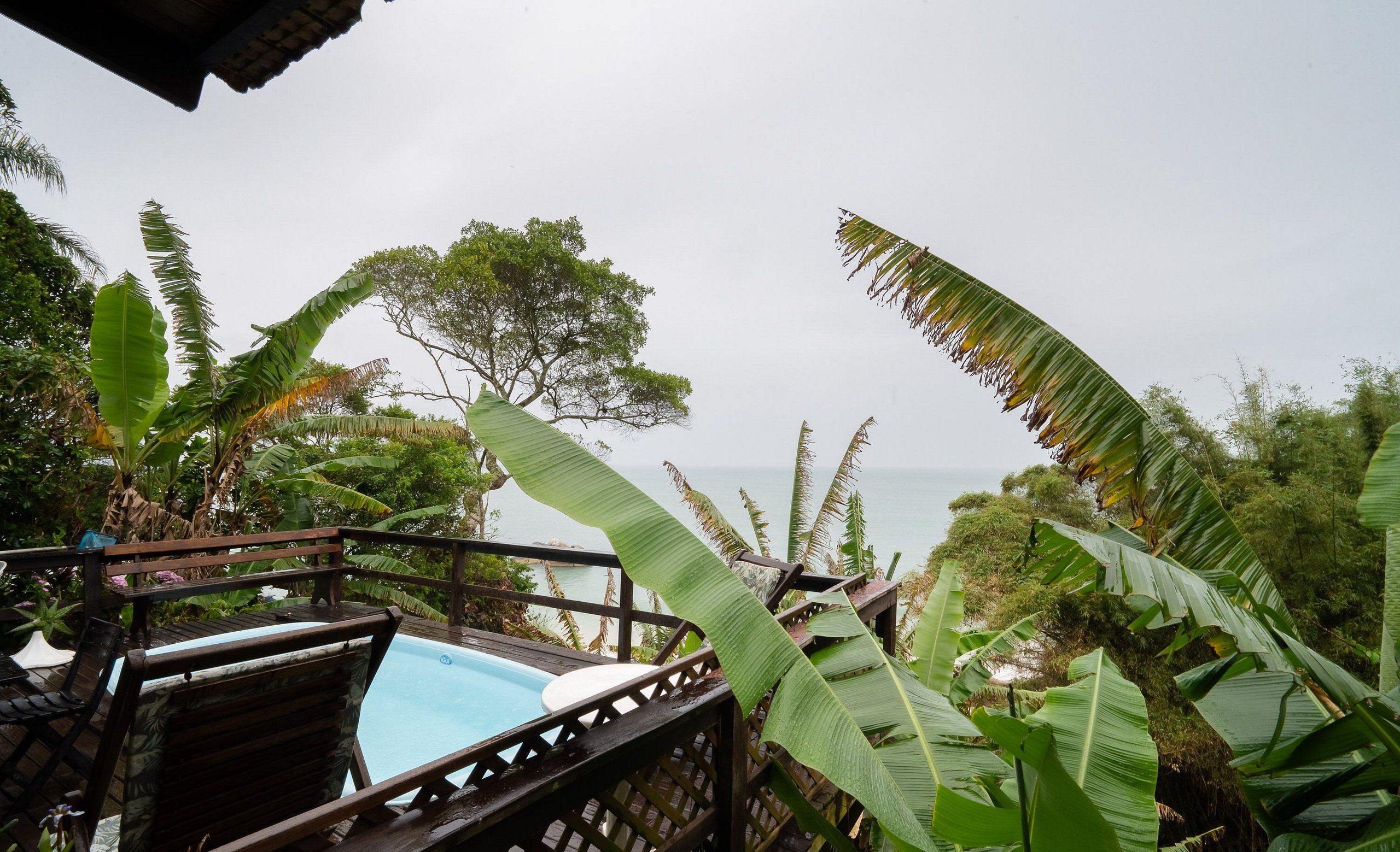 A pool area with wooden deck and lounge chairs, surrounded by tropical foliage and overlooking a body of water.