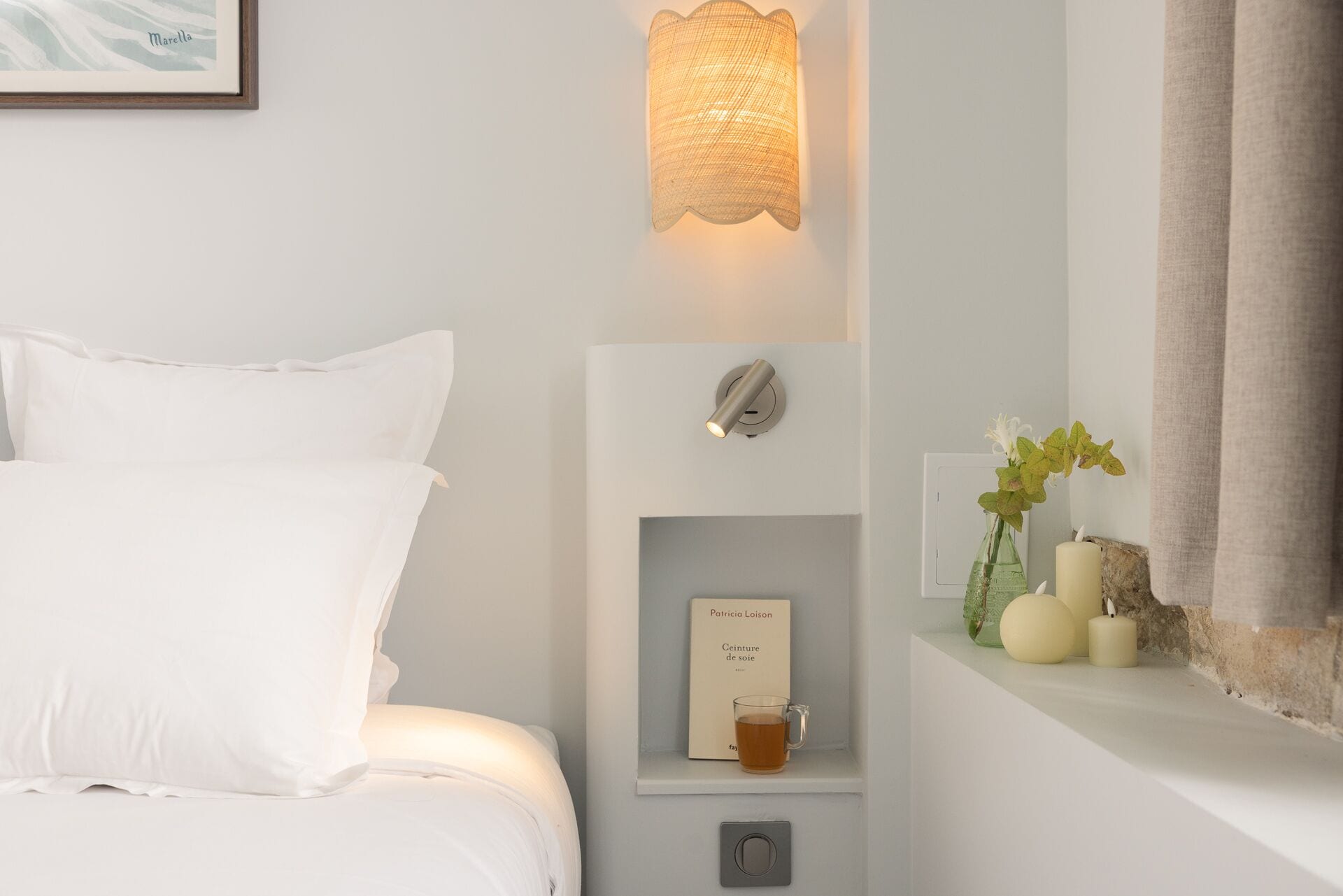 Un letto rifatto con lenzuola bianche, una lampada fissata al muro, un piccolo ripiano con un libro, una tazza di vetro e delle candele.