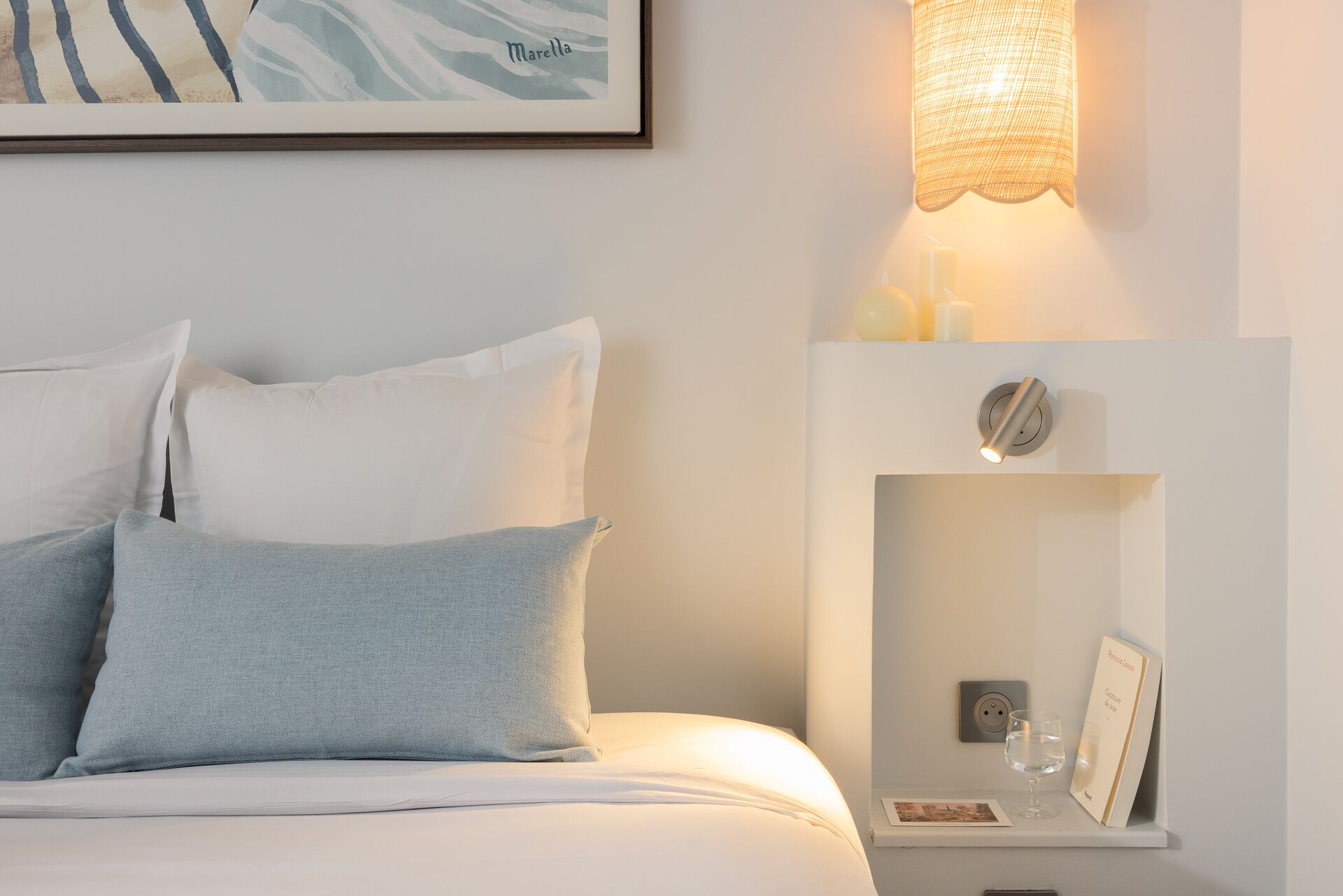 Un lit bien fait, avec des oreillers blancs et bleu clair, une lampe fixée au mur et une petite étagère sur laquelle se trouvent un verre d’eau et un cadre avec une photo.