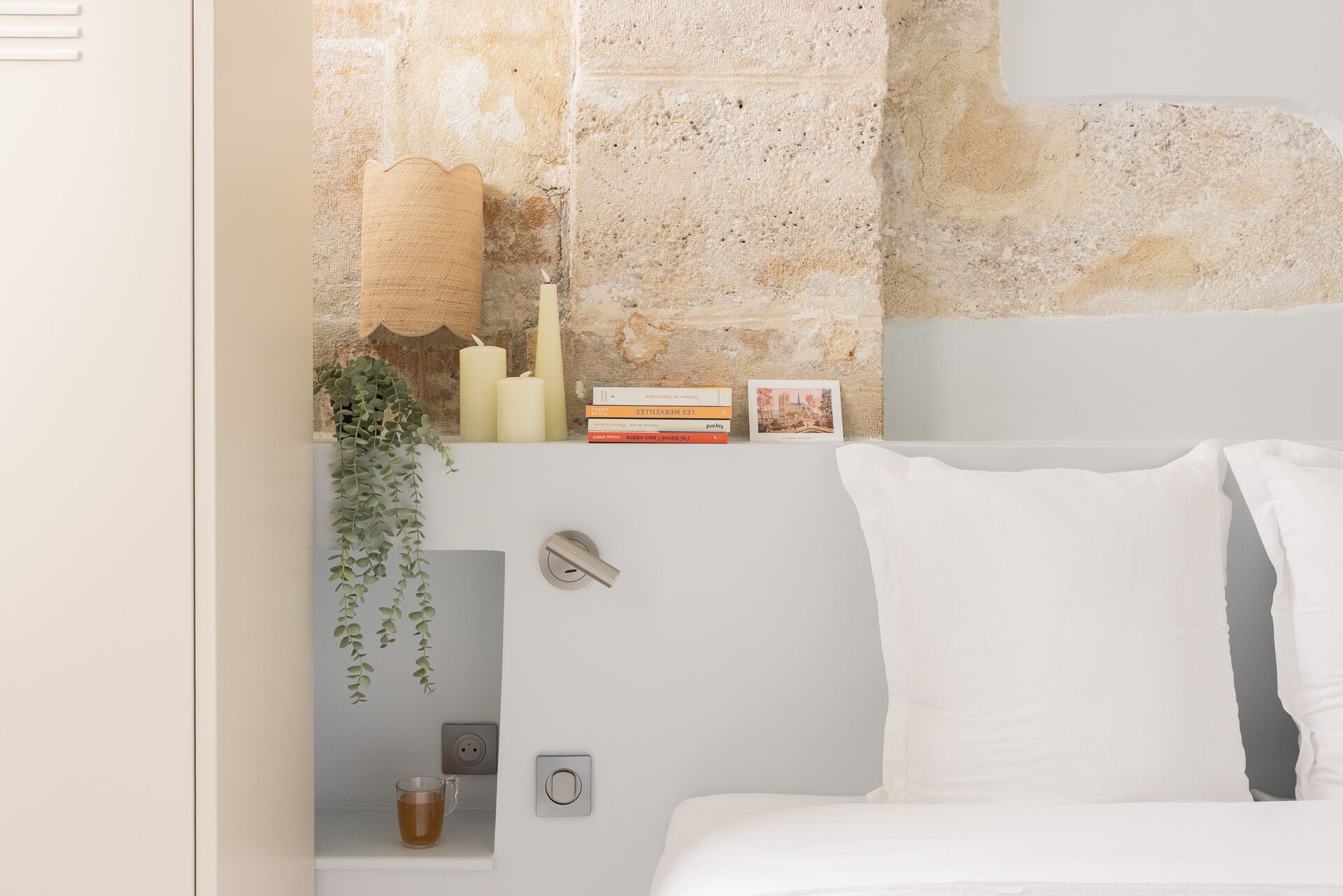 A bedroom with a bed, white pillows, a bedside table with a glass of tea, a wall-mounted outlet, and a shelf with candles, books, and a plant.