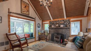 A cozy living room with a stone fireplace, wooden beams, a rocking chair, and a large window with blinds. - Third night free on stays before 8/30/26 (Government Camp)